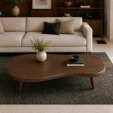 A modern wavy wooden coffee table in a warm medium brown finish, styled in a living room setting on a dark textured rug in front of a cream-colored sofa.
