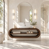 A floating wooden bathroom vanity with an integrated open shelf, holding a white vessel sink, set in a bright, modern bathroom with an arched mirror and wall sconces.